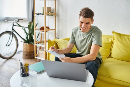 Lächelnder Typ in seinen Zwanzigern auf gelbem Sofa zu Hause bei der Kursarbeit mit Notizen und Laptop auf dem Tisch