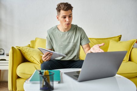 Verwirrter junger Student auf gelbem Sofa zu Hause bei der Kursarbeit in Notizen und Laptop auf Couchtisch