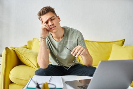 hombre cansado en el sofá amarillo con el ordenador portátil en la cara de la cubierta de la mesa de café con la mano y sosteniendo los vasos