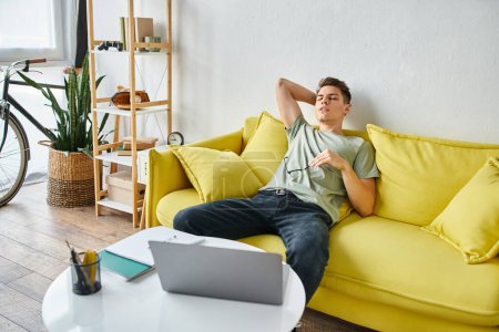 gars fatigué avec ordinateur portable mettant la main derrière la tête et se penchant sur le canapé jaune avec des lunettes