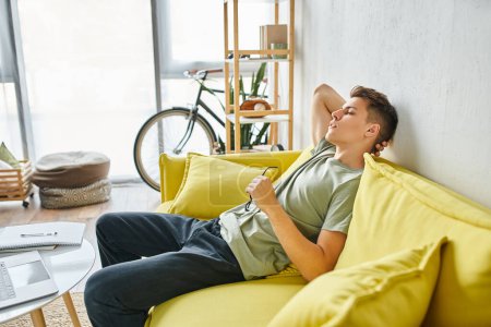 Photo for Sideways of weary man in his 20s putting hand behind head and leaning on yellow couch with glasses - Royalty Free Image