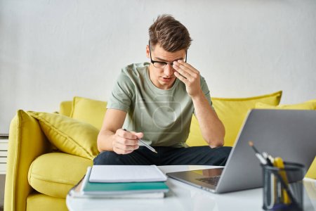 hombre atractivo cansado en sus años 20 con gafas de visión en el sofá amarillo en la red del hogar en el ordenador portátil