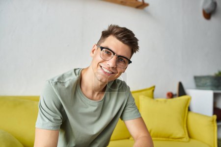 cheerful student in his 20s with vision glasses smiling on yellow couch in living room
