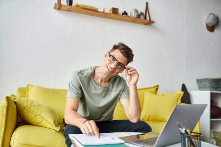 smiling man in his 20s with vision glasses on yellow couch putting pen down on coffee table