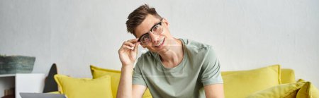 Photo for Banner of smiling man in 20s with vision glasses on yellow couch putting pen down on coffee table - Royalty Free Image