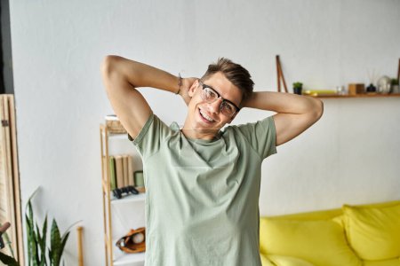 attraktiver Kerl mit braunen Haaren und Brille im Wohnzimmer, der die Arme hinter dem Kopf vor die Kamera legt