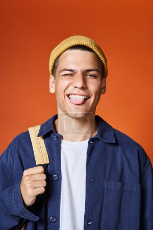 verspielter junger Mann mit gelbem Hut und Rucksack, der seine Zunge auf Terrakottahintergrund ausstreckt