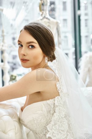 A young brunette bride in a flowing white wedding dress, seated gracefully on a luxurious couch in a bridal salon.