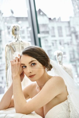 Eine junge brünette Braut in einem fließenden weißen Brautkleid lehnt anmutig auf einem luxuriösen Bett in einem Brautsalon.