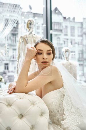 Photo for A young brunette bride in a wedding dress gracefully sits on a elegant couch in a bridal salon. - Royalty Free Image