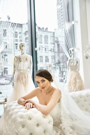 Eine junge brünette Braut im Brautkleid sitzt auf einer Bank vor einem Fenster.