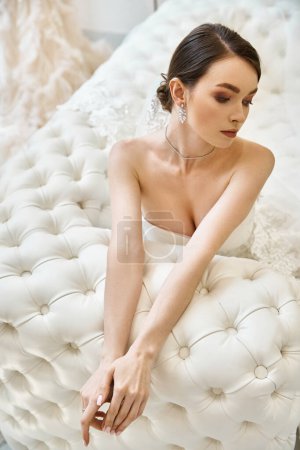 A young brunette bride in a pristine white dress sits elegantly on a plush bed in a bridal salon, radiating timeless beauty.