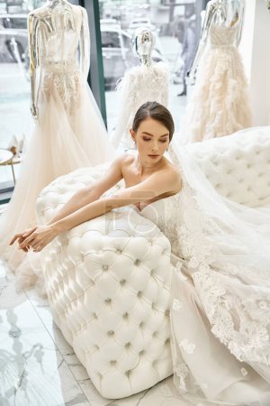 A young brunette bride in a wedding dress sits gracefully on top of a white couch, exuding elegance and poise.