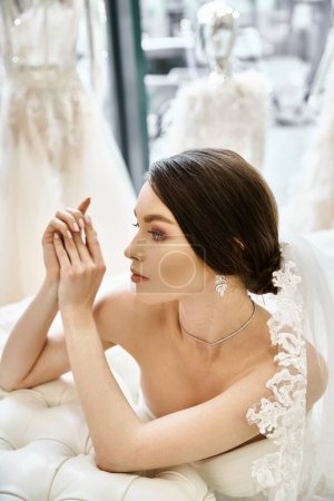 A young brunette bride in a wedding dress sits gracefully on a luxurious couch in a bridal salon.