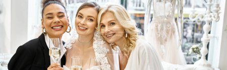Photo for A young brunette bride in a wedding dress and her middle-aged blonde mother in a bridal salon, standing proudly together. - Royalty Free Image