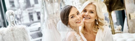 Téléchargez les photos : Une jeune mariée brune dans une robe de mariée et sa mère blonde d'âge moyen se tenant côte à côte dans un salon de mariée. - en image libre de droit
