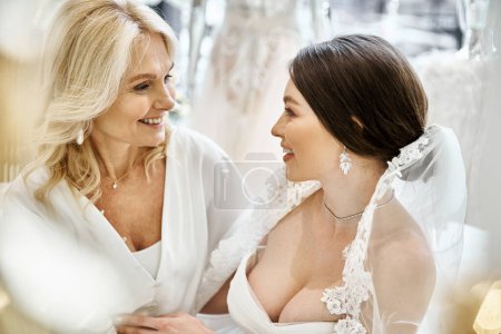 Eine junge brünette Braut im Brautkleid steht neben ihrer blonden Mutter mittleren Alters in einem Brautsalon.