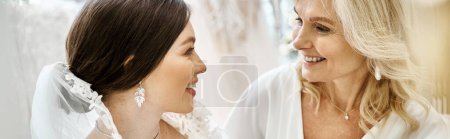 Téléchargez les photos : Une jeune mariée dans une robe de mariée se tient avec sa mère d'âge moyen dans un salon de mariée. - en image libre de droit