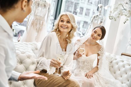 Eine junge brünette Braut im Brautkleid, ihre Brautjungfer und eine blonde Mutter mittleren Alters sitzen in einem Brautsalon zusammen.
