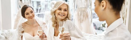 A young bride in a wedding dress raises her champagne flute with her two bridesmaids in a bridal salon.