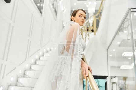 Téléchargez les photos : Une jeune mariée brune, vêtue d'une robe de mariée, debout gracieusement sur un escalier dans un salon de mariée. - en image libre de droit