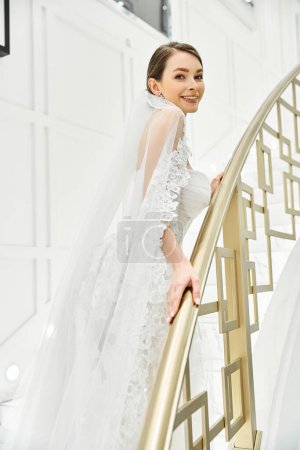 A young brunette bride in a wedding dress stands gracefully on a staircase in a bridal salon.