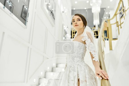 Foto de Una joven novia morena en un vestido de novia se para en una escalera en un salón de novias. - Imagen libre de derechos
