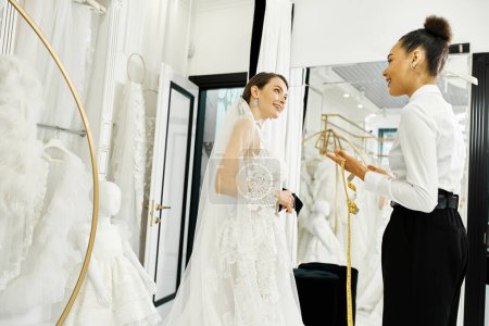 Eine junge brünette Braut im Hochzeitskleid, die mit einer afroamerikanischen Shopping-Assistentin in einem Brautsalon vor einem Spiegel steht.