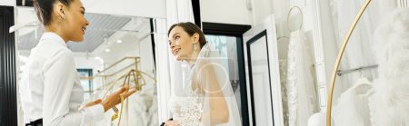 A young brunette bride in a wedding dress and her shopping assistant standing in front of a mirror in a bridal salon.