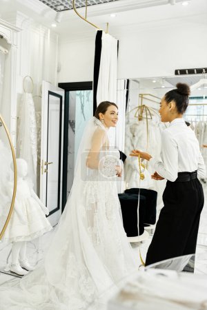 A young bride in a white dress and assistant stand together, gazing at their reflections in a mirror in a bridal salon.