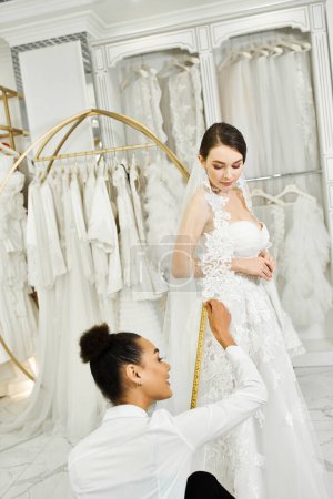 A young brunette bride in a wedding dress is being measured by an African American shopping assistant in a bridal salon.