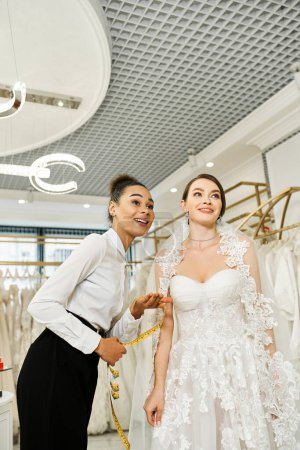 Young brunette bride in a white wedding dress standing next to an African American woman in a black dress in a bridal salon.