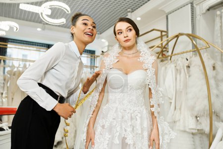 A young brunette bride in a wedding dress stands next to an African American shopping assistant in a bridal salon.
