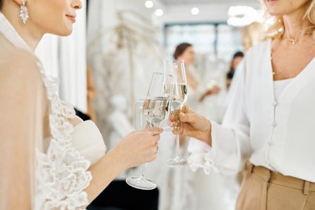 A young brunette bride in a wedding dress stands with her middle-aged mother, both holding champagne glasses in a bridal salon.