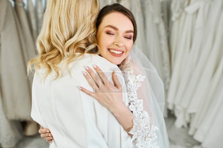 A young bride in a wedding dress lovingly embraces her middle-aged mother in a bridal salon.