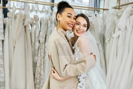 A young bride in a white wedding dress embracing her bridesmaid in front of a rack of various wedding dresses in a bridal salon.
