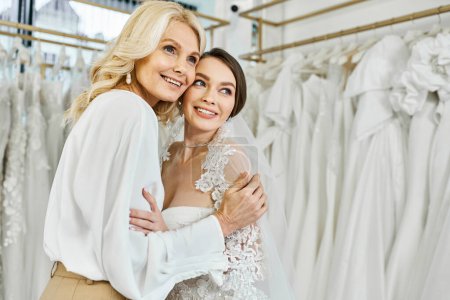 A young brunette bride in a wedding dress embraces her middle-aged mother in a bridal salon filled with wedding dresses.