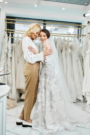 A young bride in a white wedding dress hugs her middle-aged mother, both surrounded by a display of elegant wedding gowns.