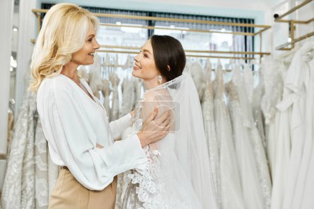 A young brunette bride and her middle-aged mother are standing next to each other in front of a rack of dresses in a bridal salon.