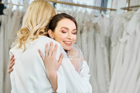 A young bride in a wedding dress hugs her middle-aged mother in a bridal salon in front of a rack of dresses.