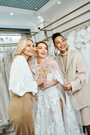 Una joven novia en un vestido de novia, su madre de mediana edad, y su mejor amiga como dama de honor de pie delante de un estante de vestidos.