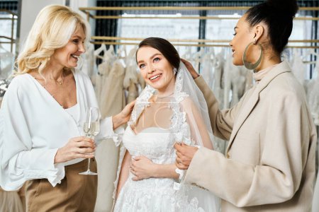 Frauen, eine Braut im weißen Hochzeitskleid, ihre Mutter und ihre beste Freundin als Brautjungfer, stehen zusammen vor einem Ständer mit bunten Kleidern.