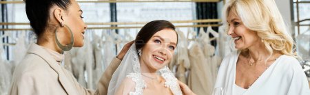 Une jeune mariée brune, sa mère d'âge moyen, et meilleure amie comme demoiselle d'honneur se tiennent près d'un rack de robes dans un salon de mariée.