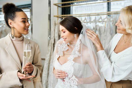 Deux femmes, une jeune mariée en robe blanche, et l'autre sa mère, se tiennent près d'une étagère de robes dans un salon de mariée.