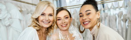 A young bride in a wedding dress, her middle-aged mother, and her best friend as a bridesmaid stand together in a bridal salon.