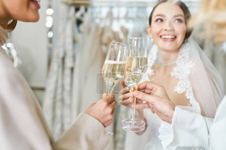 Young bride, her mother, and best friend standing in a bridal salon, raising glasses of champagne in a celebratory toast.