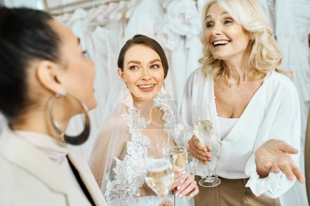 Foto de Una joven novia con un vestido de novia, su madre de mediana edad, y una dama de honor en un salón de novias, sosteniendo copas de vino. - Imagen libre de derechos