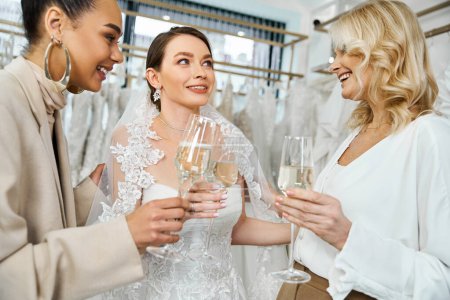 Photo for A young brunette bride in a wedding dress, her middle-aged mother, and best friend as a bridesmaid, standing together, holding wine glasses. - Royalty Free Image