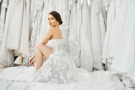 Eine junge brünette Braut sitzt vor einem Kleiderständer und wählt sorgfältig das perfekte Kleid für ihren Hochzeitstag aus.