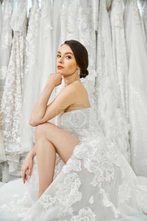 Eine junge brünette Braut in einem weißen Kleid sitzt nachdenklich vor einem Kleiderständer in einem Hochzeitssalon.